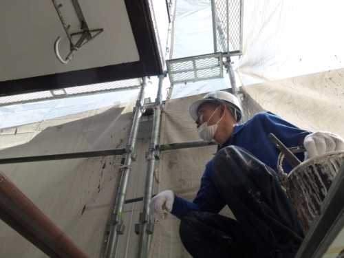 koushin_P1030158-1 | 外壁塗装は神奈川県横浜市の塗装職人 Construction worker in a blue uniform and hard hat crouches on scaffolding, wearing a mask and gloves outside a building.