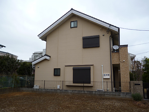 Two-story beige house with dark shutters, a small attic window, satellite dish, brick entry column, and a fenced yard under an overcast sky.