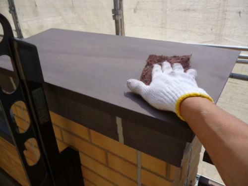 Gloved hand smoothing a brown patch of filler onto a dark metal sheet outdoors on scaffolding above a brick wall.