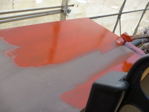 Close‑up of a red paint being brushed onto a gray surface, with a paint brush applying a glossy coat in a workshop setting.