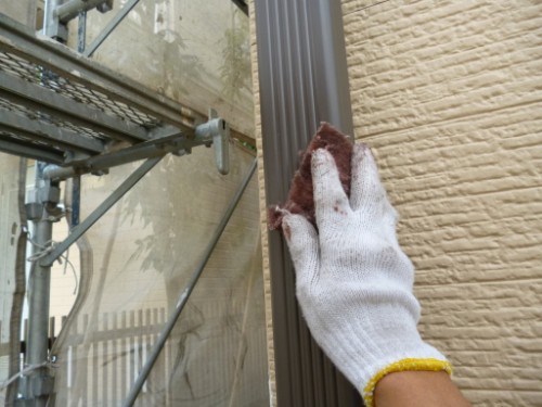 Gloved hand scrubbing a brown metal door frame beside scaffolding on a beige brick wall.