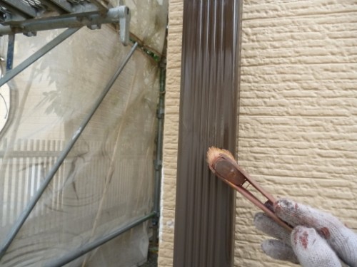 Gloved hand applies brown paint to a vertical metal trim beside a beige brick wall using a brush.