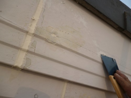 Close-up of a hand with a blue putty knife smoothing beige sealant along horizontal vinyl siding on an exterior wall.