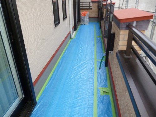 Narrow balcony walkway covered with blue protective flooring, edged by green tape, with railing on the right and windows on the left.