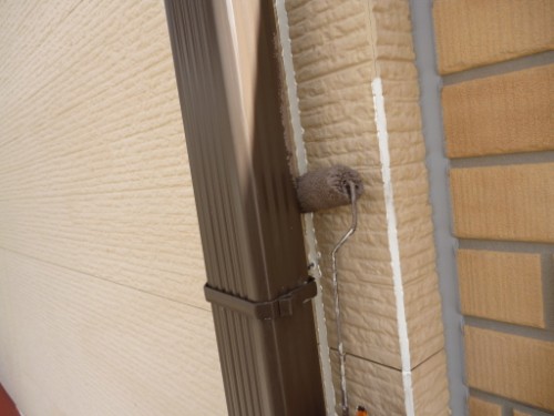 Close-up of a paint roller applying white sealant along a vertical seam between beige textured wall and brick, with a brown rain gutter to the left.