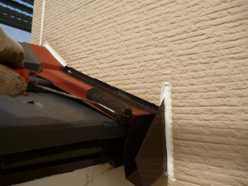 Gloved hand using an orange caulk gun to apply sealant at the joint between a dark gutter and a beige brick wall exterior.