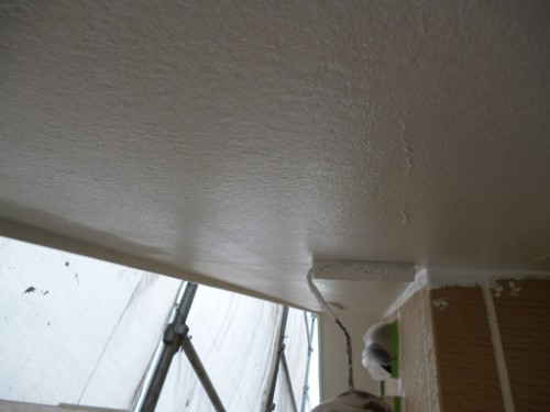 Painter applying white paint to a ceiling with a roller; paint drips along the wall seam, with scaffolding visible outside the window.