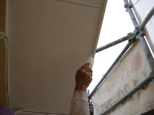 Hands applying plaster with a trowel to a ceiling edge, scaffolding visible in the background.