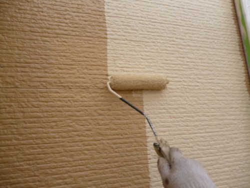 Hand in a white glove uses a paint roller to apply tan/beige paint to a textured exterior wall with a two-tone finish.