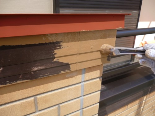 Gloved hand painting a beige brick exterior with dark brown paint using a brush, with orange trim above.