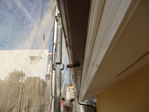 Construction worker wearing a white glove holds a tool along the edge of a beige building, with scaffold and blue plastic sheeting in the background.