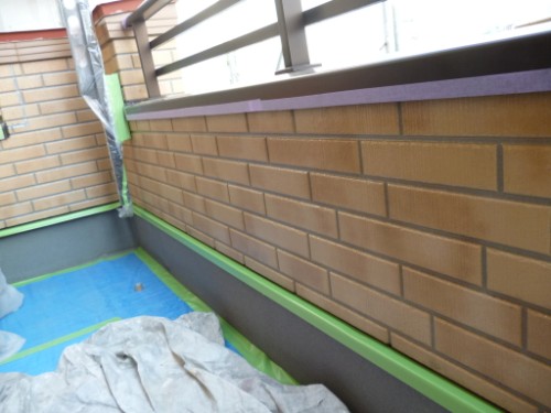 Balcony edge under renovation: brown brick wall with green painter's tape along the base and blue protective tarp on the floor with some cloths/polish rags nearby.