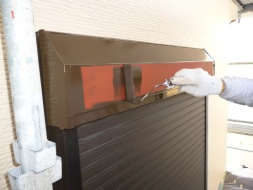 Person wearing a glove uses a paint roller to apply brown paint to the top edge of a metal outdoor box/shutter against a beige wall.