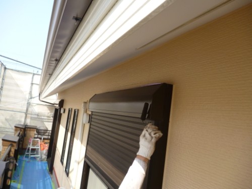 Worker's gloved hand adjusting a brown exterior window shutter on a beige wall, with scaffolding nearby in a construction site setting.
