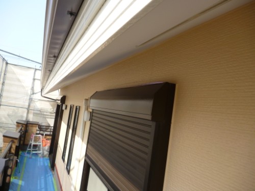 Exterior view of a house with a brown rolling shutter over a window, beige textured wall, white eaves, and scaffolding with a ladder in the background.