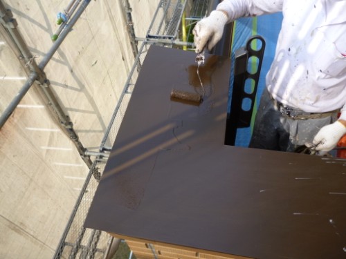 Worker wearing gloves applies dark brown paint to a metal panel on scaffolding outdoors, using a roller.