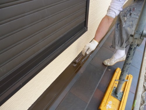 Painter on scaffolding applying brown paint along the base of a beige wall with dark brown trim, wearing gloves and paint-splattered clothes.