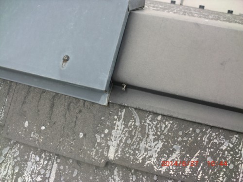 Close-up of a metal roof seam with gray flashing, showing chipped paint, a small screw, and white weathering streaks.
