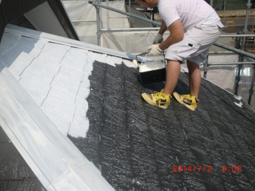 Worker on a sloped roof applying a white coating with a brush while standing on scaffolding.