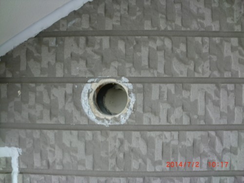 Circular pipe opening in textured beige exterior siding with white sealant around the edge.