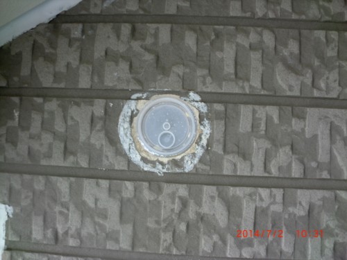 Round metal access cap (likely a sewer cleanout) set in a textured exterior wall with white crust around its edge.
