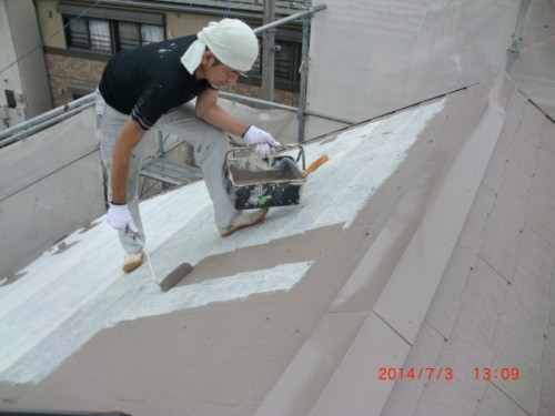 Worker on a sloped roof applying light gray paint with a roller and tray near scaffolding.