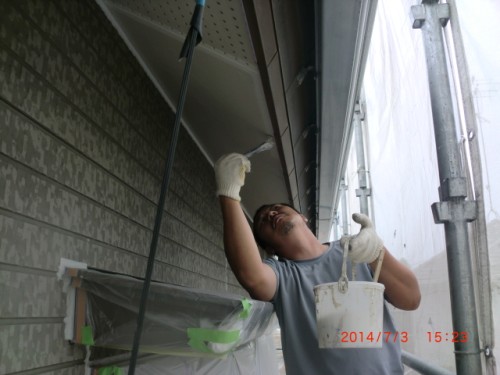 Man wearing gloves on scaffolding, painting the exterior of a house with a brush and holding a paint bucket.
