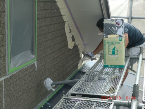 Worker on scaffolding applying patch material to a brick exterior, masking tape around a window; toolbox on the platform.