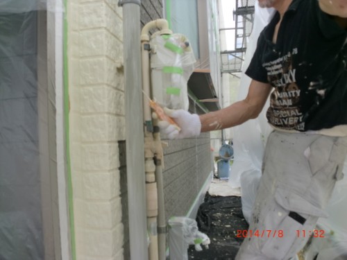 Construction worker in gloves applies masking tape around exterior gas pipes on a building during renovation/painting work.