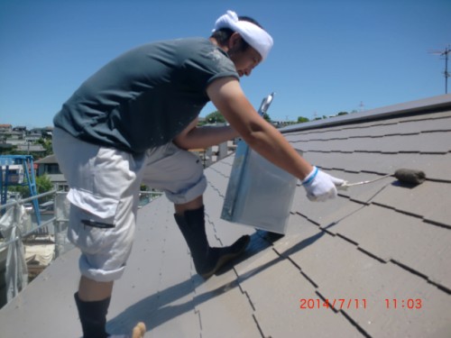 Person on a sloped roof, kneeling and applying coating with a roller while wearing gloves and a white head covering.