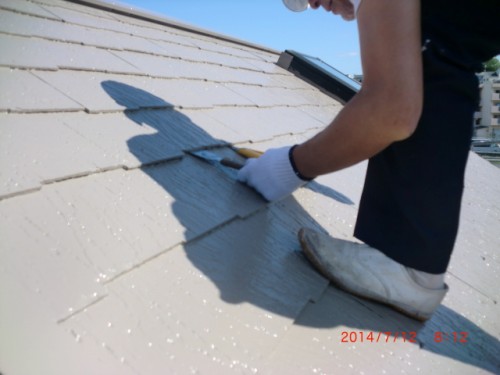 Worker kneels on a sloped roof, applying gray sealant with a trowel; wears white gloves and boots, with a tool resting nearby.
