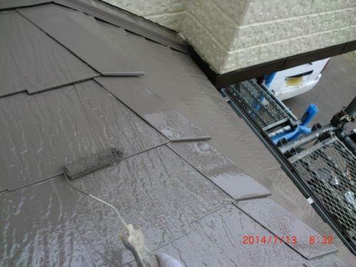 Close-up of a person painting brown coating on roof shingles with a roller; scaffold and building wall visible.