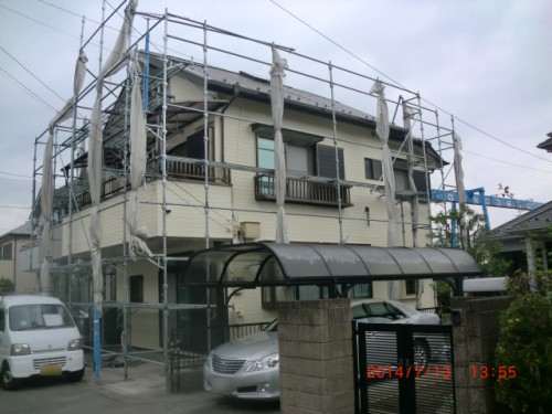 Two-story beige house under metal scaffolding with protective netting, a curved carport, and several parked cars in the driveway.