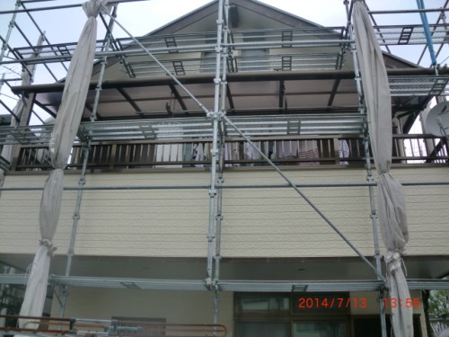Beige two-story house wrapped in metal scaffolding with fabric coverings; laundry hanging on the balcony.