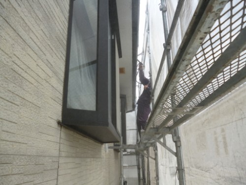 koushin_P1050774-1 | 外壁塗装は神奈川県横浜市の塗装職人 Worker in purple clothing on narrow scaffold between two building walls, performing window work at height.