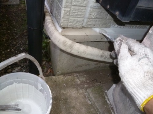 koushin_P1050838-1 | 外壁塗装は神奈川県横浜市の塗装職人 Gloved hand holding a beige flexible hose near the building exterior, with a white bucket on the ground nearby.