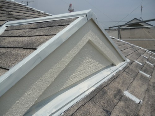 koushin_P1050844-1 | 外壁塗装は神奈川県横浜市の塗装職人 Close-up of a residential roof ridge with white flashing at the peak and beige textured gable, surrounded by dark gray shingles.