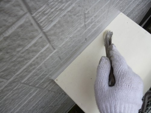 koushin_P1050905-1 | 外壁塗装は神奈川県横浜市の塗装職人 Gloved hand holding a trowel and applying plaster to a white panel mounted on a grey brick wall.