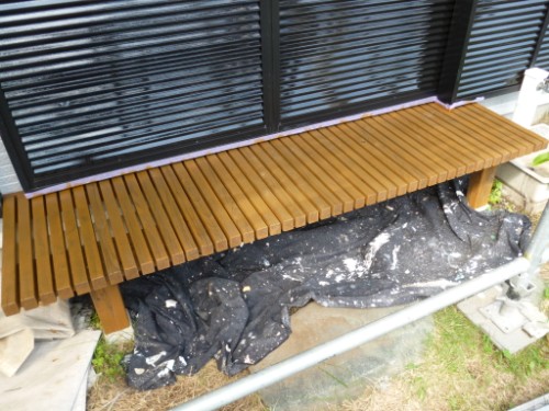 koushin_P1050914-1 | 外壁塗装は神奈川県横浜市の塗装職人 Long brown wooden slatted bench installed outside against a wall with black horizontal blinds above it.