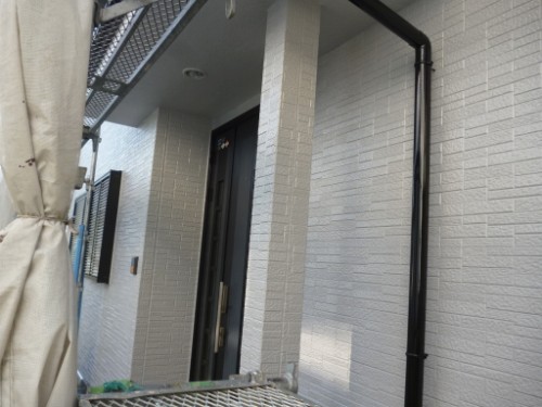 Exterior view of a light-gray brick building entrance under construction, with scaffolding on the left and a black rain downspout along the wall.