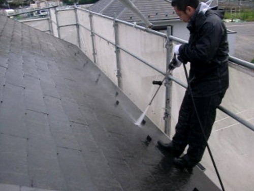 A man wearing dark clothing uses a long wand-like tool to wash a sloped roof with soapy water near a metal railing.