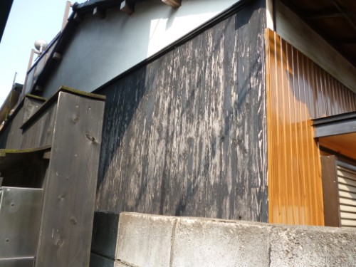 Weathered black wooden exterior wall with orange vertical trim and a concrete base at a corner of a building under a sloped roof.
