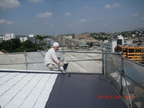 Construction worker in white safety gear sits on a rooftop edge with a laptop, surrounded by scaffolding and a city skyline in the background.