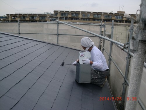 Worker in white clothing kneels on a dark sloped roof, operating a circular-dial gauge mounted in a metal box on a workday scene in the cityscape.
