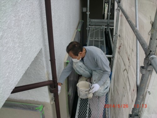 Worker wearing a mask and gloves on scaffolding, painting an exterior white wall with a paint bucket in hand.