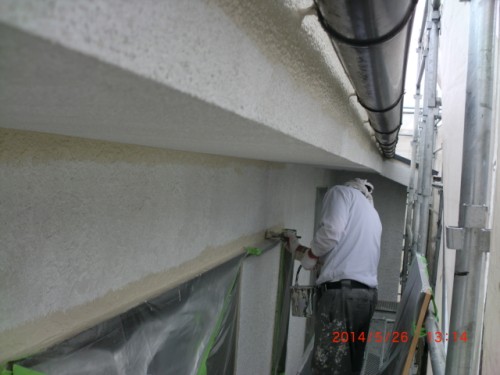 Worker on scaffolding painting a beige stucco exterior wall; a person in a hooded jacket using a roller, with construction tape and equipment visible, date stamp 2014/05/26.