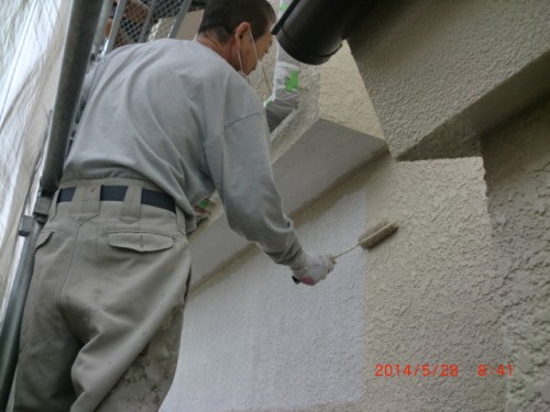 A man in a mask and gloves paints an exterior stucco wall with a roller while standing on scaffolding.