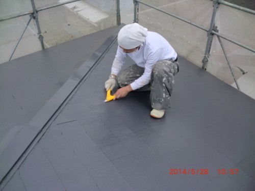 Roofer kneels on a gray flat roof, smoothing the surface with a yellow trowel, wearing white clothing and gloves, scaffolding around.