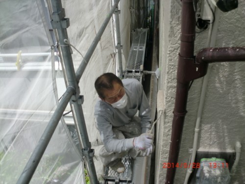 Construction worker in white coveralls and a mask kneels on scaffolding, applying coating to an exterior wall beside a pipe.
