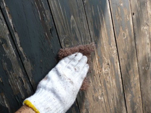Gloved hand scrubbing a weathered wooden fence with a brown abrasive pad.